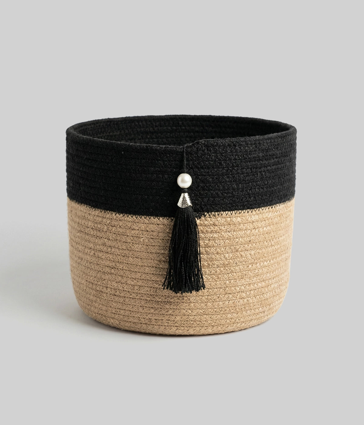 The Black Tassel Basket is a round, woven سلة تخزين featuring a black upper half, natural tan lower half, and accented with a black tassel and two silver beads on the front, presented against a plain gray background.