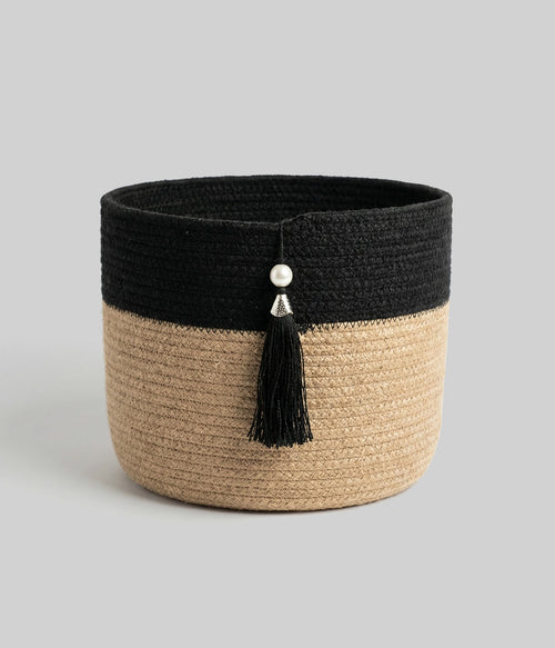 The Black Tassel Basket is a round, woven سلة تخزين featuring a black upper half, natural tan lower half, and accented with a black tassel and two silver beads on the front, presented against a plain gray background.