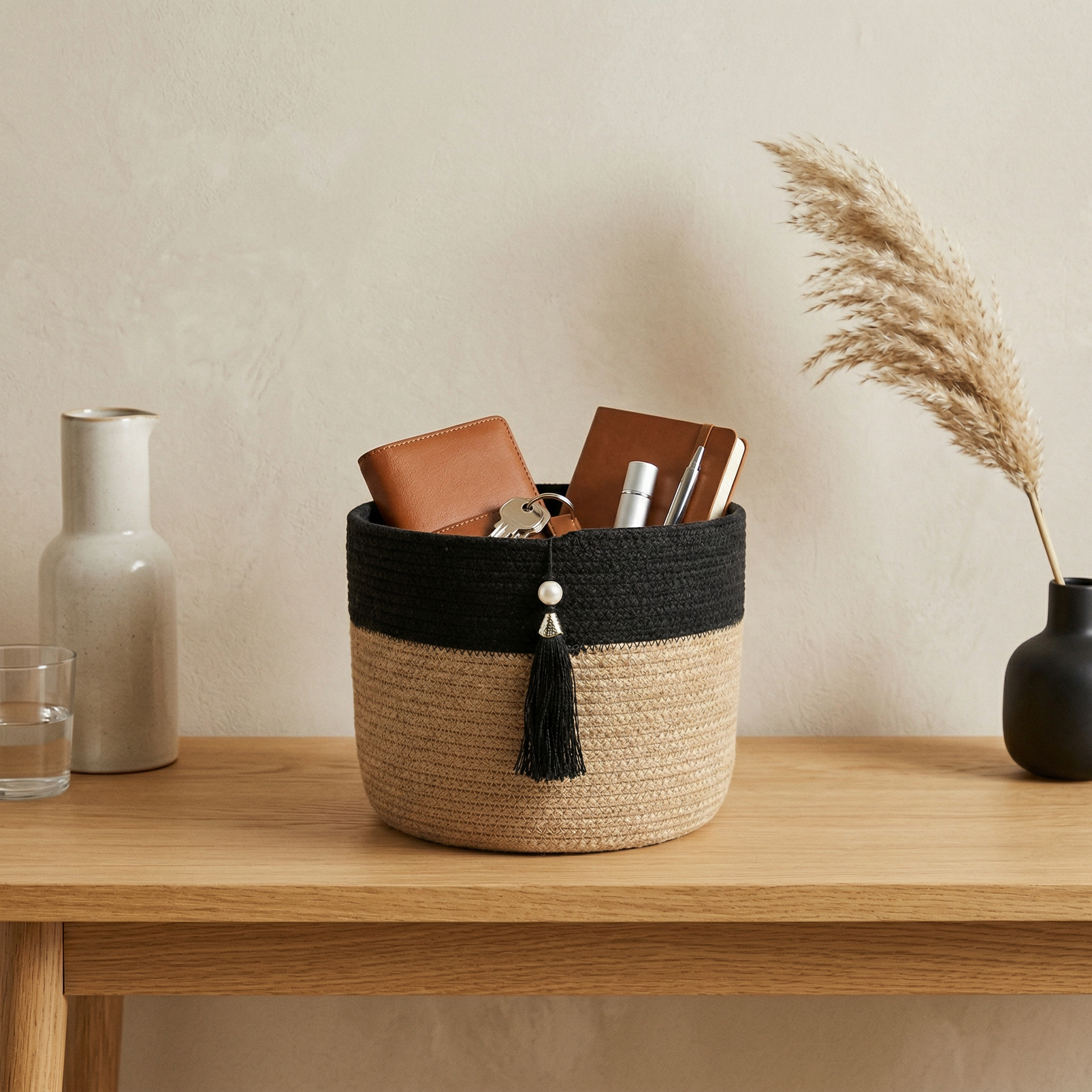 تجلس Black Tassel Basket بغطائها الأسود وشرابتها على طاولة خشبية، تحتوي على محفظة بنية ودفتر وقلم ومفاتيح. بجوارها مزهرية سيراميك بها ماء، مزهرية بيج طويلة وسلة ديكور سوداء صغيرة مع عشب مجفف.
