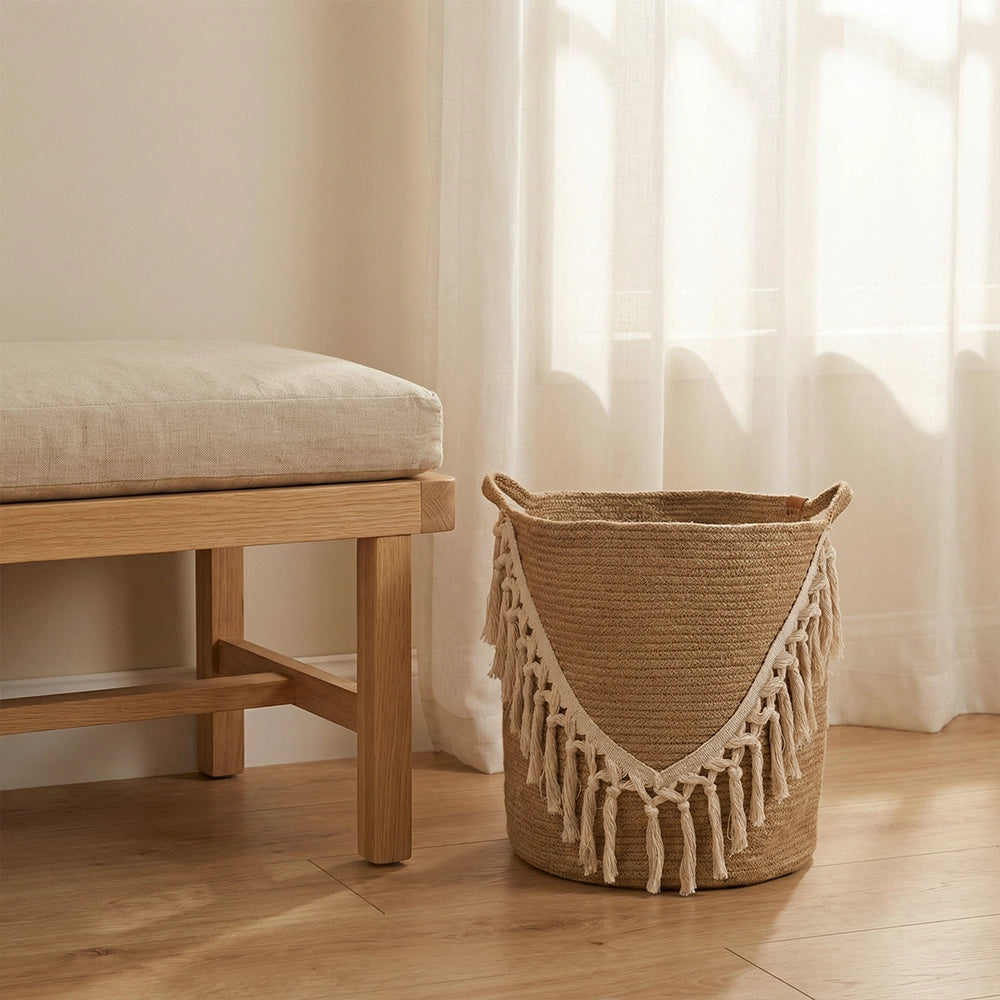 سلة Bohème بتفاصيل شرابة بيضاء موضوعة على أرضية خشبية بجانب مقعد فاتح مع وسادة بيج، وتضفي الستائر الشفافة في الخلفية لمسة من الإشراق والتنظيم الأنيق والدافئ للمساحة.