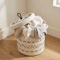 The Geo Basket, a woven cotton storage basket with black geometric patterns and a partially open lid, overflows with white and gray clothes on a wooden floor near a sunlit window.