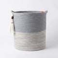 The Ocean Gray Basket is a round woven باسكت يدوي قطني with two handles, featuring a light gray bottom and dark gray top. A decorative tassel with beads hangs from one handle. This باسكت ديكور is displayed on a plain white background.