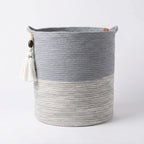 The Ocean Gray Basket is a round woven باسكت يدوي قطني with two handles, featuring a light gray bottom and dark gray top. A decorative tassel with beads hangs from one handle. This باسكت ديكور is displayed on a plain white background.