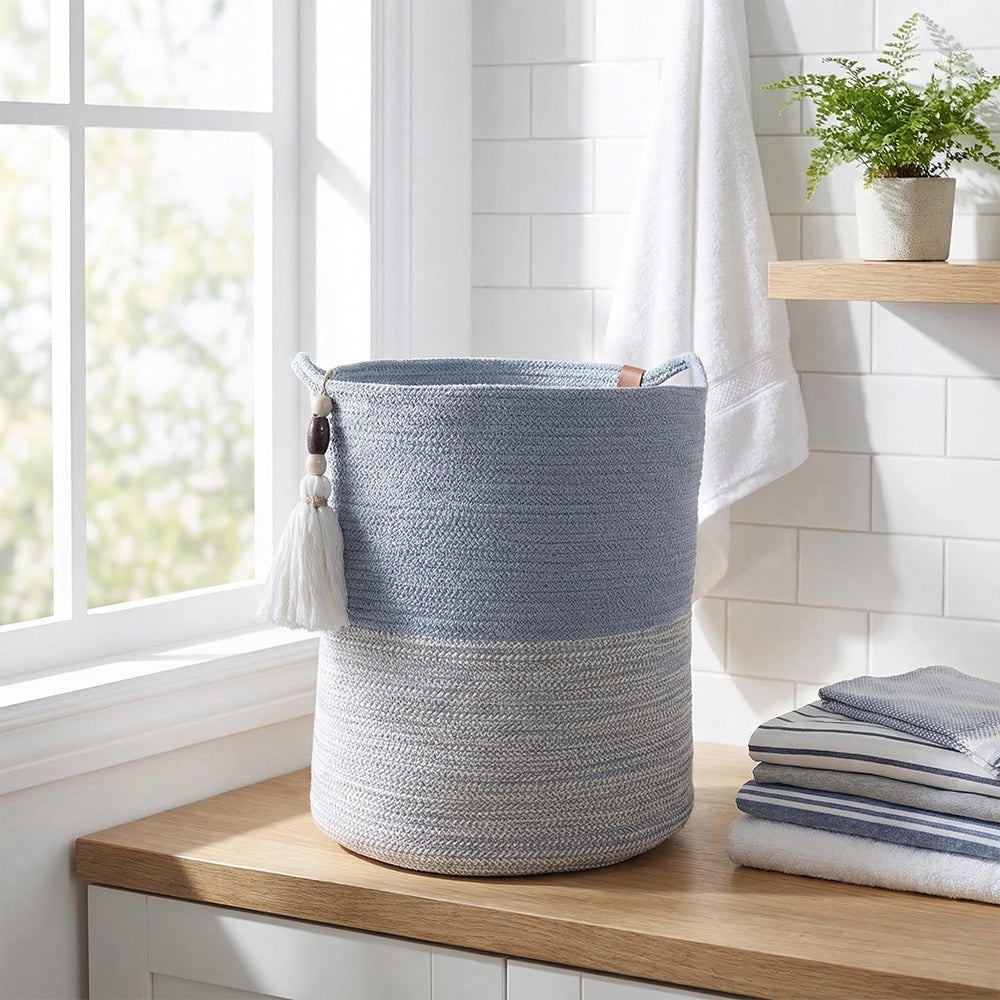 An Ocean Gray Basket with decorative tassels sits on a wooden countertop in a bright bathroom near a window, next to folded towels and a small potted fern.