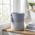 An Ocean Gray Basket with decorative tassels sits on a wooden countertop in a bright bathroom near a window, next to folded towels and a small potted fern.