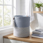 An Ocean Gray Basket with decorative tassels sits on a wooden countertop in a bright bathroom near a window, next to folded towels and a small potted fern.