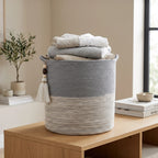An Ocean Gray Basket with a tassel and neutral tones sits on a wooden table, filled with neatly folded clothes. Nearby, a potted plant, stacked books, and a bowl rest by a sunlit window.