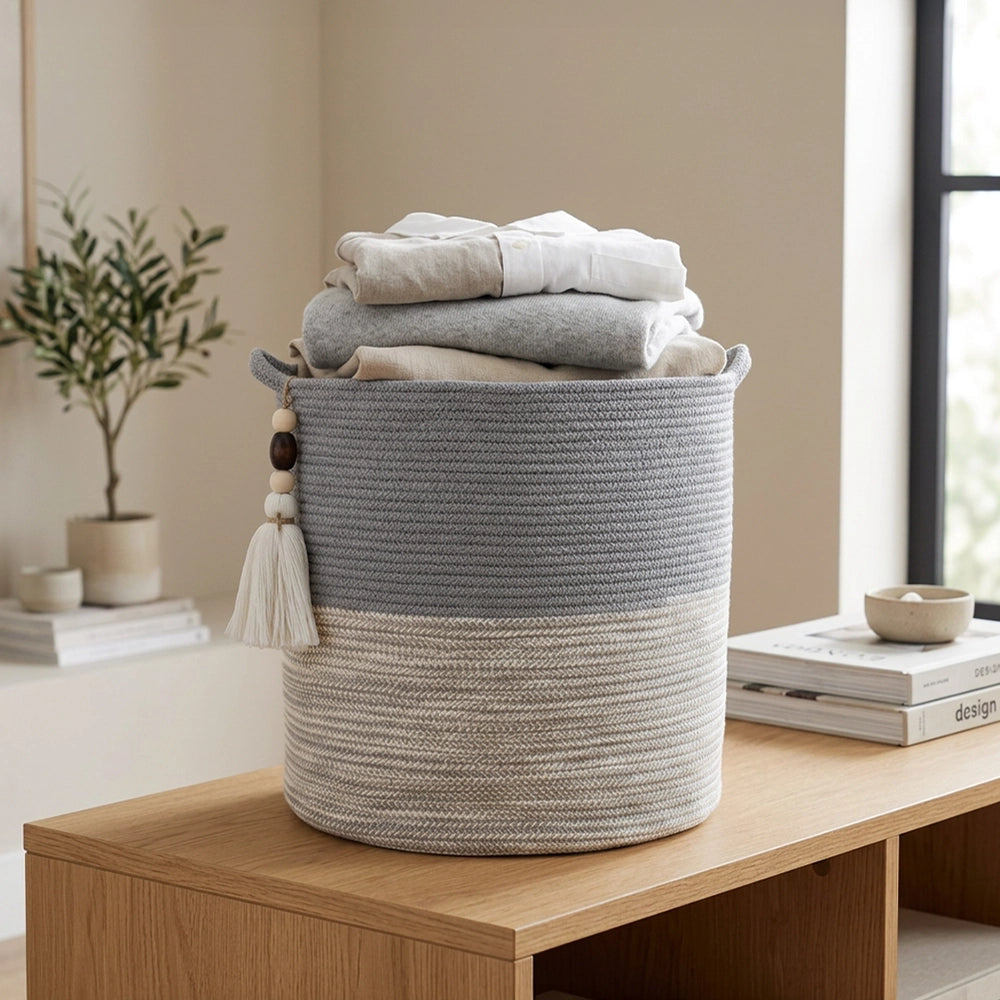 An Ocean Gray Basket with a tassel and neutral tones sits on a wooden table, filled with neatly folded clothes. Nearby, a potted plant, stacked books, and a bowl rest by a sunlit window.