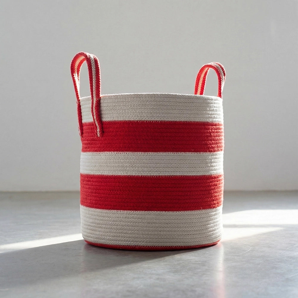 The Terra Red Basket, a round cotton storage basket with two handles and red-and-white horizontal stripes, sits on a smooth light surface with sunlight casting a soft shadow.