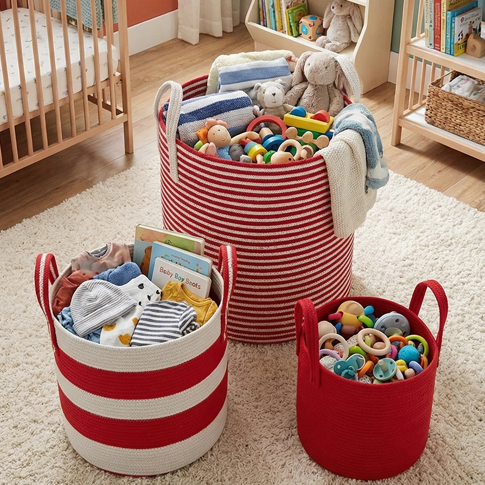 Three Terra Red Baskets (باسكت تخزين قطني) with red and white stripes sit on a white rug, holding baby essentials—plush toys, rattles, books, and clothes—in a cozy nursery with a crib and shelves in the background.